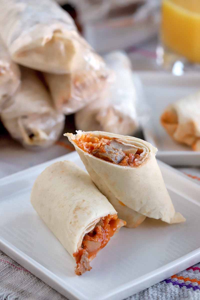 A breakfast burrito sliced in half at an angle and arranged on a square white ceramic plate, with more wrapped in plastic and stacked in the background, with a glass of orange juice in soft focus.