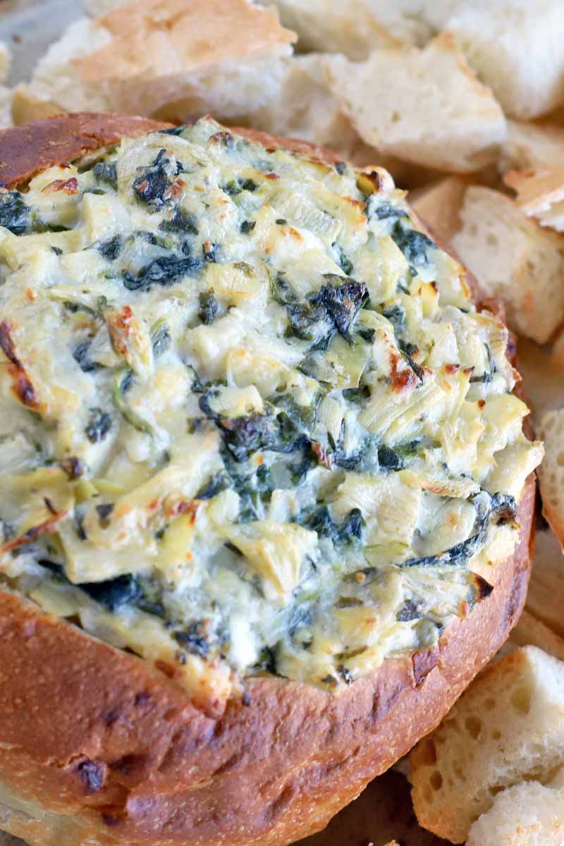 Closeup vertical shot of a boule loaf filled with homemade spinach artichoke dip baked to have a golden brown crust on top, with chunks of bread for serving in the background.