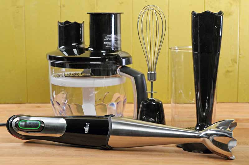 The Braun MQ777 Multiquick Hand Blender Set with accessories on a maple wooden table and green wooden background.