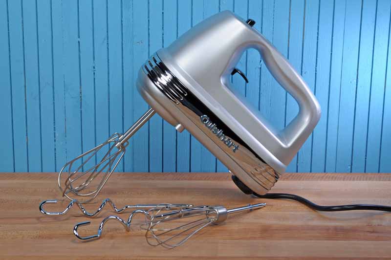 The Cuisinart HM-90BSC Power Advantage Plus 9 Speed Hand Mixer sitting on a maple wooden butcher block counter with a bead board background painted a medium blue.