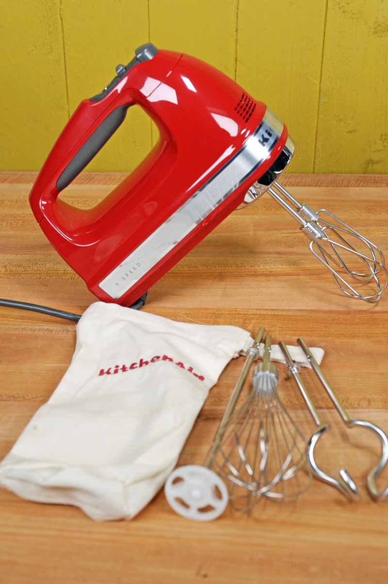 An Empire Red version of the KitchenAid KHM926ER 9-Speed Hand Mixer with attachments, cloth storage bag on maple butcher block table.