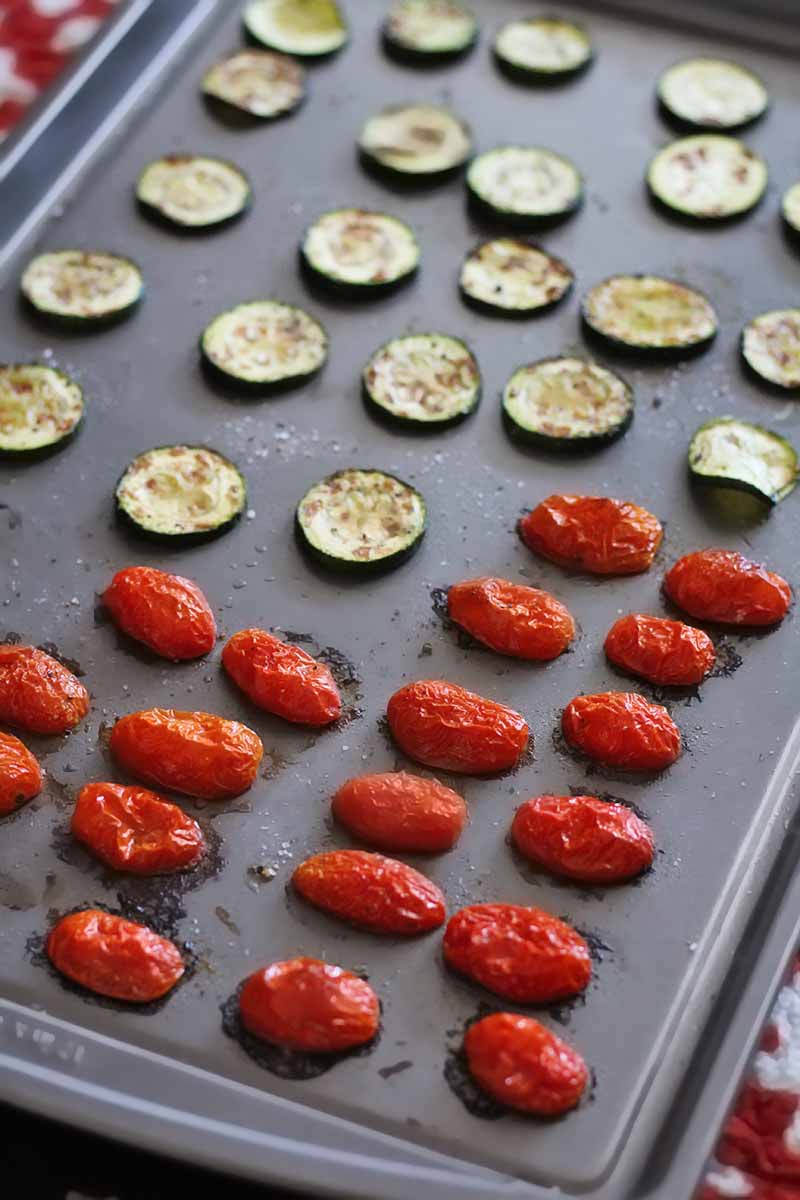 Overhead oblique vertical shot of roasted halved grape tomatoes and slices of zucchini on a metal rimmed baking pan.