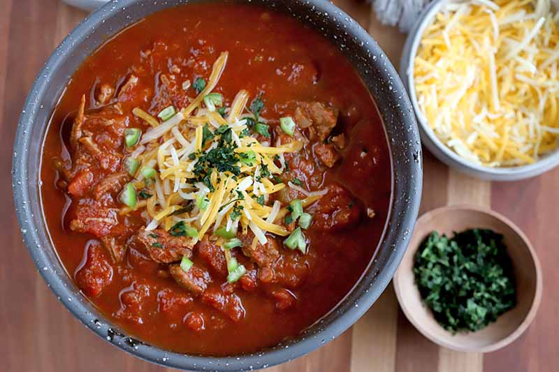 Overhead shot of a bowl of chili with smaller bowls of shredded cheese and chopped fresh cilantro, on a striped light brown wood surface.