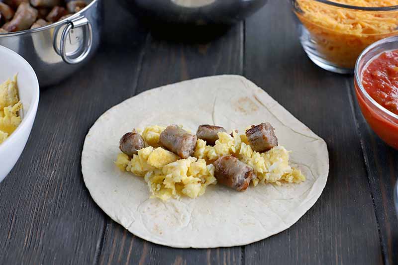 Sliced cooked sausage and scrambled egg are arranged at the center of a flour tortilla, on a brown wood table with bowls of egg, meat, cheese, and salsa around the edge of the frame.