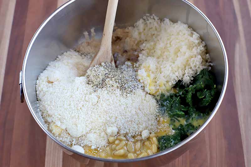 A stainless steel mixing bowl of cheese, egg, pine nuts, spinach, garlic, onion, salt, and pepper, with a wooden spoon, on a striped brown and beige wood surface.