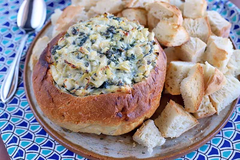 A hollowed boule stuffed with homemade spinach artichoke dip with a golden brown crust on top, next to chunks of bread for serving on a white and terra cotta ceramic plate, with a spoon on a dark and light blue patterned cloth surface.