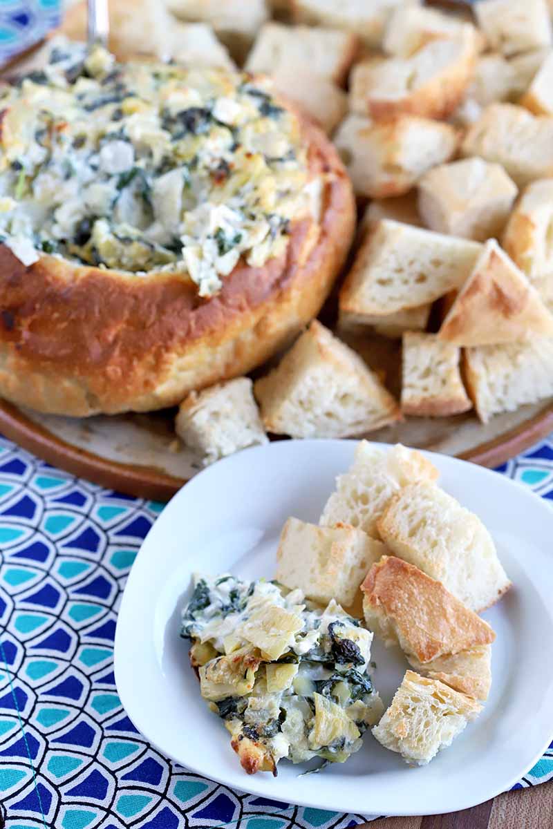 Vertical oblique shot of a serving platter and a plate of spinach artichoke dip with chunks of bread for serving, on a dark and light blue patterned cloth.