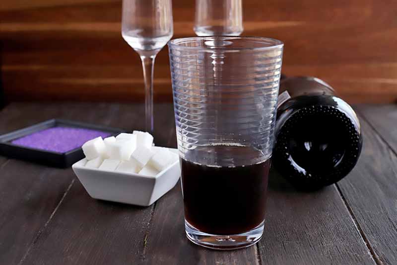 A pint glass is about 1/3 full of a brown liquid, with a square white dish of white cubes and a square black dish of purple sanding sugar, two pieces of tall and narrow glass stemware, and a bottle of champagne on its side, on a brown wood table with a lighter brown background.