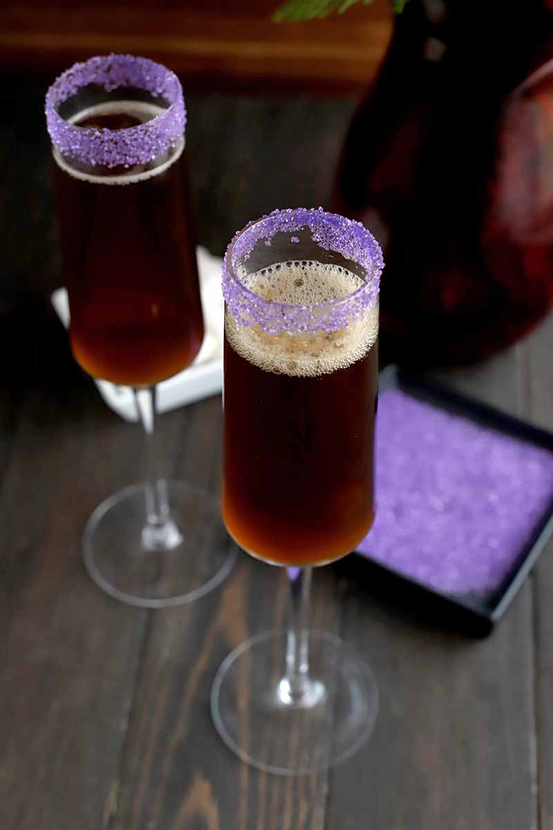 Vertical oblique shot of two champagne flutes filled with a brown beverage with purple sugar rims, with a black square dish of sanding sugar and a white square dish of sugar cubes beside a bottle of champagne on a dark brown wood surface, with a lighter brown background.
