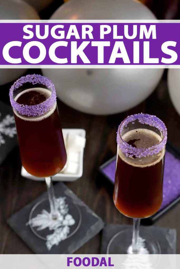 Vertical oblique shot of two champagne flutes with purple sugar rims, filled with a brown beverage, on a dark brown wood surface with slate coasters and gray balloons, a white square dish of sugar cubes, and a black square dish of purple sanding sugar.