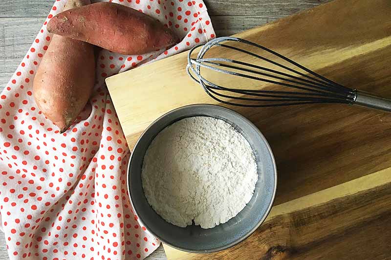Horizontal image of a gray bowl with a flour mixture on a cutting board next to a whisk.