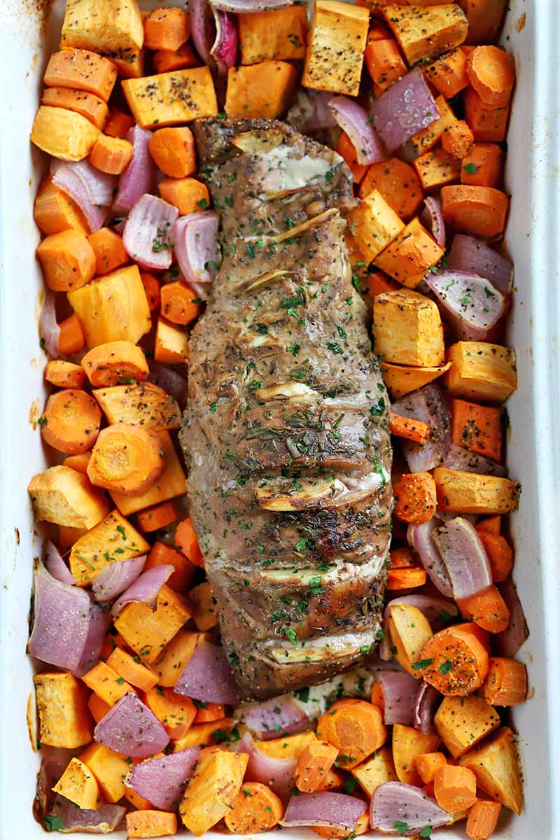 Overhead vertical shot of roasted pork tenderloin with roasted sweet potato, carrot, and red onion, in a white rectangular ceramic baking dish.