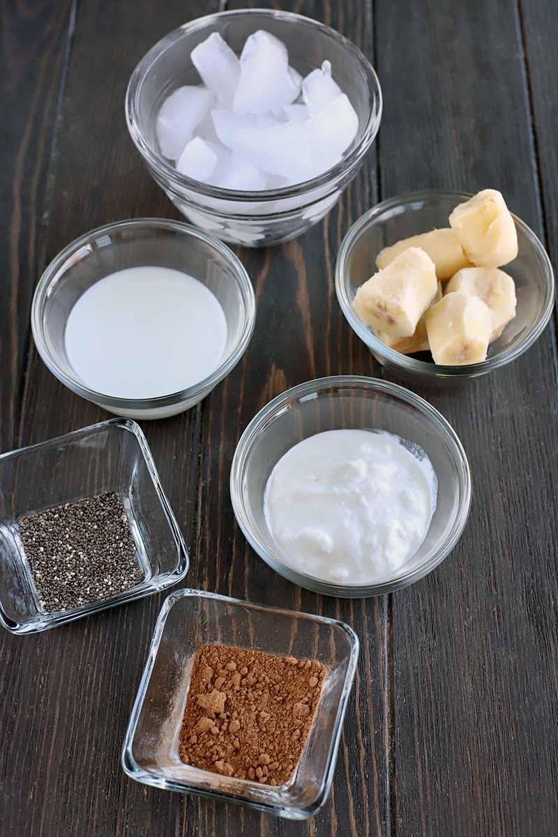 Four round and two square glass dishes of ice, sliced banana, milk, yogurt, chia seeds, and cocoa powder, on a dark brown wood table.