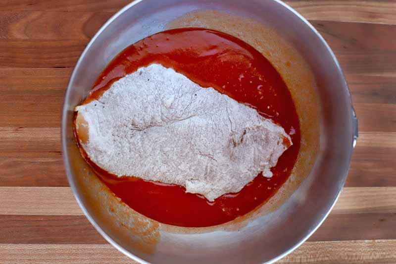 A piece of raw chicken breast dredged in flour is being dipped in a butter and hot sauce mixture in a stainless steel bowl, on a striped brown wood table.