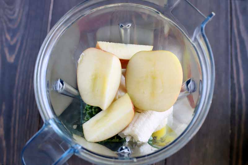 Apple, banana, and kale in a clear plastic blender pitcher, on a dark brown wood surface.
