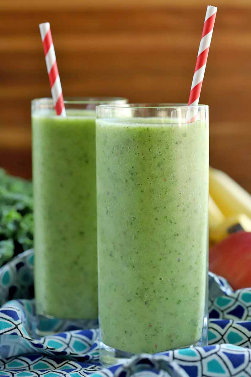 Two tall glasses of green homemade smoothie with red and white striped straws, on a dark and light blue patterned cloth with apples, bananas, and kale in the background, against a brown horizontally striped backdrop.