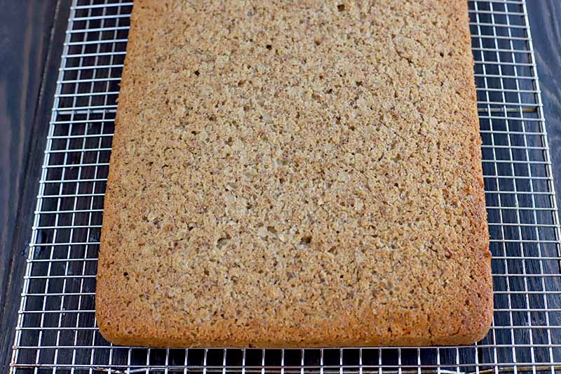 A whole slab of freshly baked gluten-free oat bars before they have been cut, on a metal cooling rack on top of a dark brown surface.