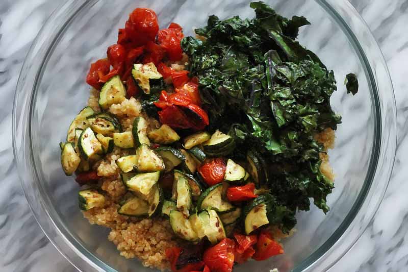 Overhead shot of piles of sauteed kale, roasted tomatoes and zucchini, and cooked quinoa in a large glass mixing bowl, on a marble surface.