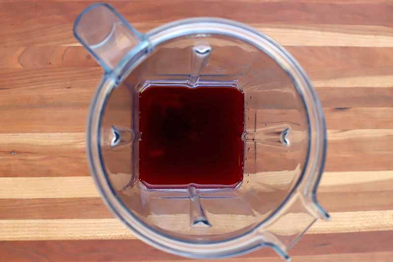 Overhead shot of a clear plastic blender pitcher with pomegranate juice at the bottom, on a brown and beige striped wood surface.