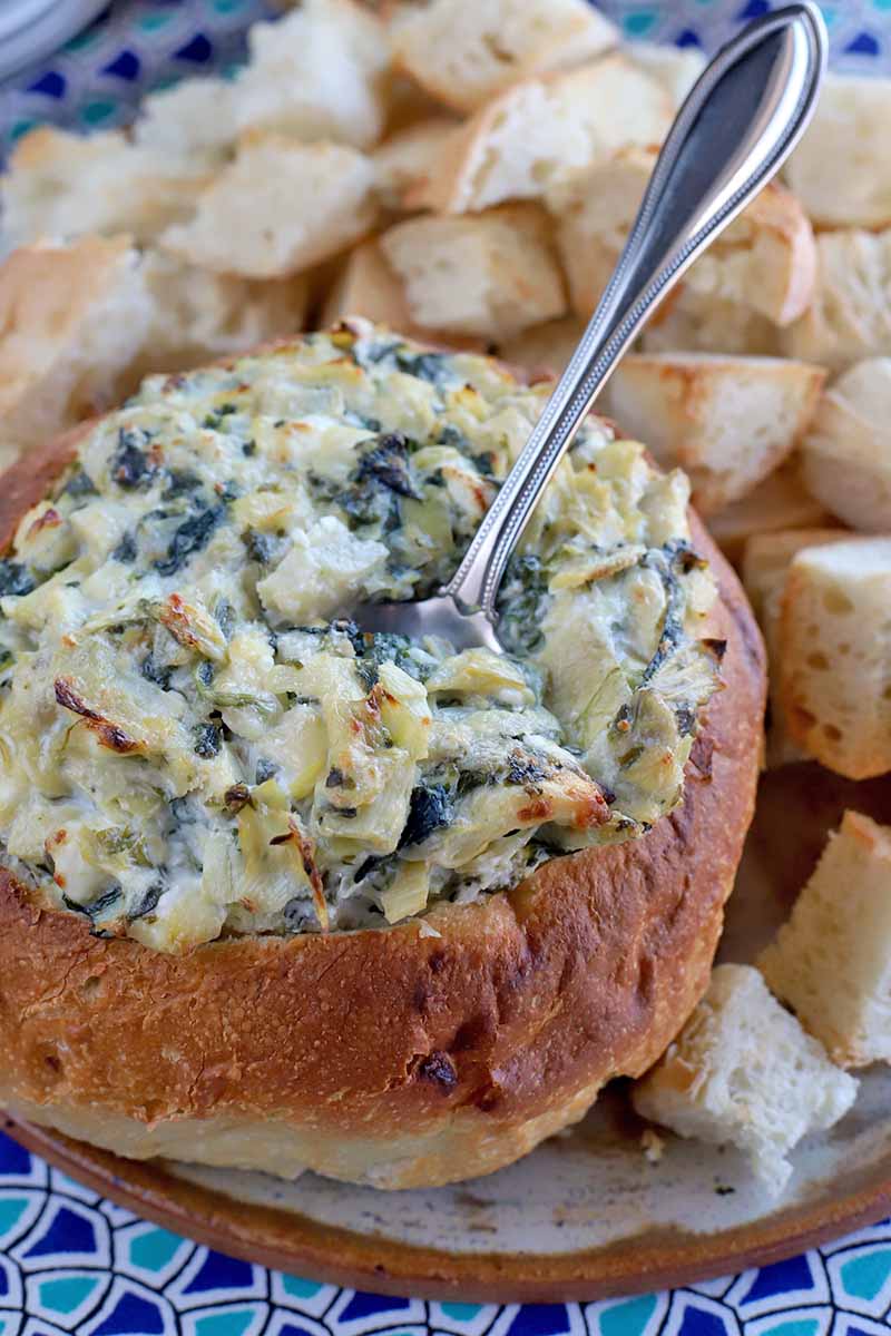 A spoon is stuck into the top of a homemade spinach and artichoke dip in a hollowed out boule loaf, with chunks of bread for serving in the background, on a white and terra cotta ceramic plate on top of a dark and light blue patterned cloth.