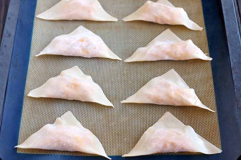 Eight unbaked wonton wrappers, filled and folded into triangles, on a silicone baking mat with a blue border, set into a rimmed metal baking sheet.