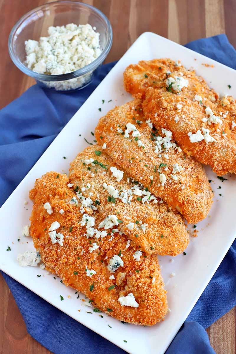 Overhead shot of four Buffalo-style chicken cutlets arranged on a rectangular serving platter and topped with cheese crumbles and fresh herbs, on top of a folded and gathered blue cloth with a small glass bowl of blue cheese to the left, on a striped brown wood table.