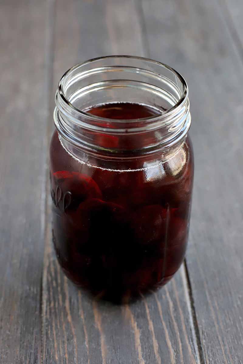 Vertical image of a glass jar filled with maraschino cherries and sherry, on a dark brown wood surface.