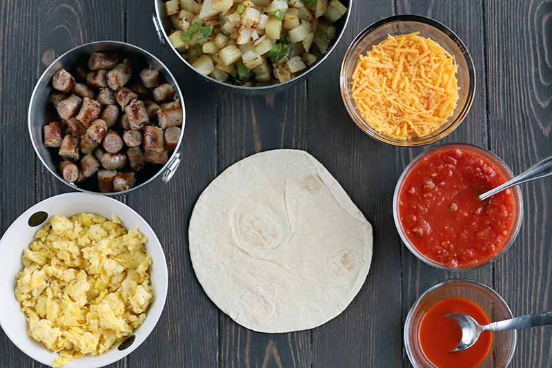 Overhead shot of a flour tortilla surrounded by bowls of scrambled egg, sliced sausage, cooked vegetables, shredded cheese, salsa, and hot sauce, with two spoons stuck into the sauces, on a dark brown wood table.