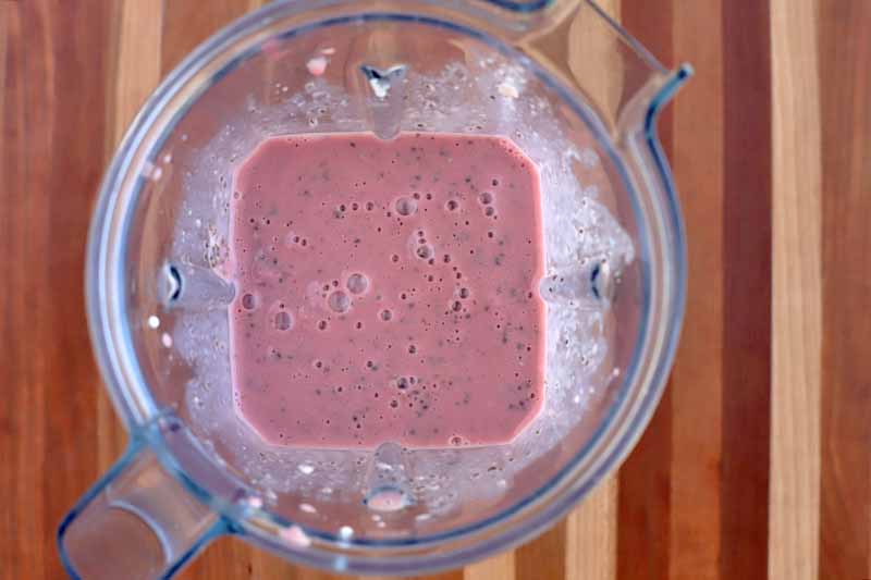 Overhead shot of a pink frothy mixture in a clear plastic blender pitcher canister, on a striped brown wood surface.