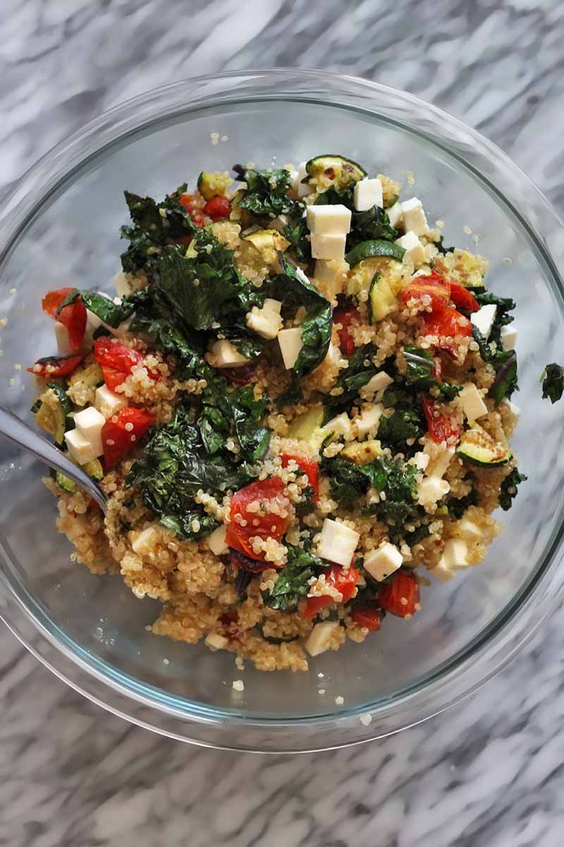 Overhead shot of a large glass mixing bowl filled with a mixture of cooked quinoa, sauteed kale, roasted zucchini and tomateos, and chopped feta cheese, with a spoon, on a marble surface.