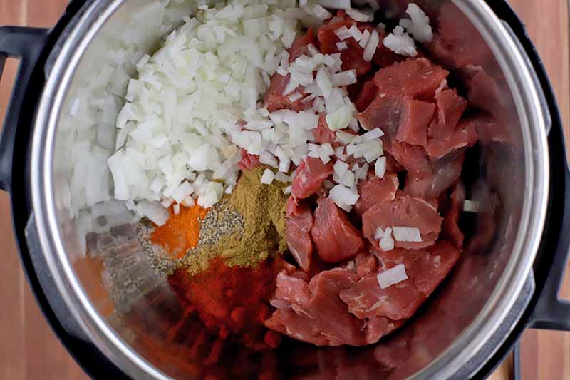 Overhead closely cropped shot of a slow cooker filled with ingredients including beef stew meat, chopped onion, and spices.
