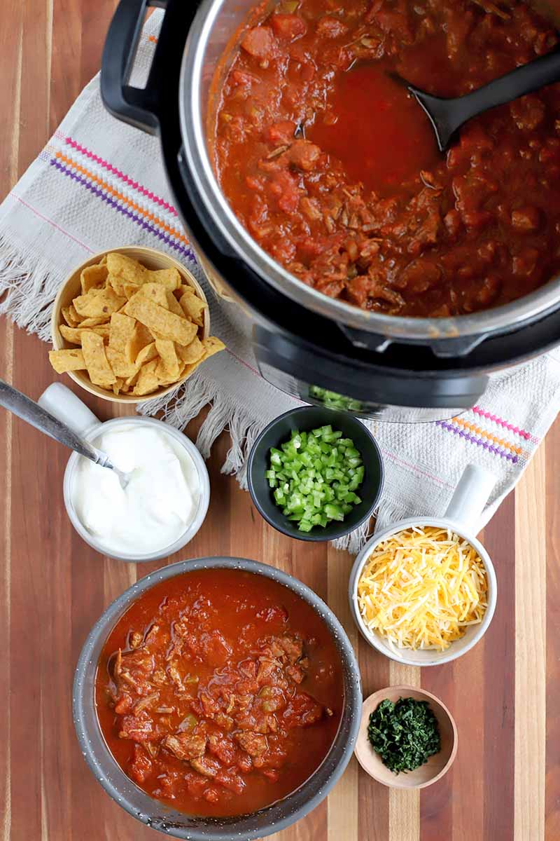 Overhead shot of a slow cooker filled with beef chili in a red sauce with a black plastic ladle, on a striped white kitchen towel with frings, next to several small bowls of toppings including corn chips, chopped green bell pepper, shredded cheese, sour cream with a spoon, and chopped cilantro, with a portion dished out in a gray bowl, on a brown striped surface.