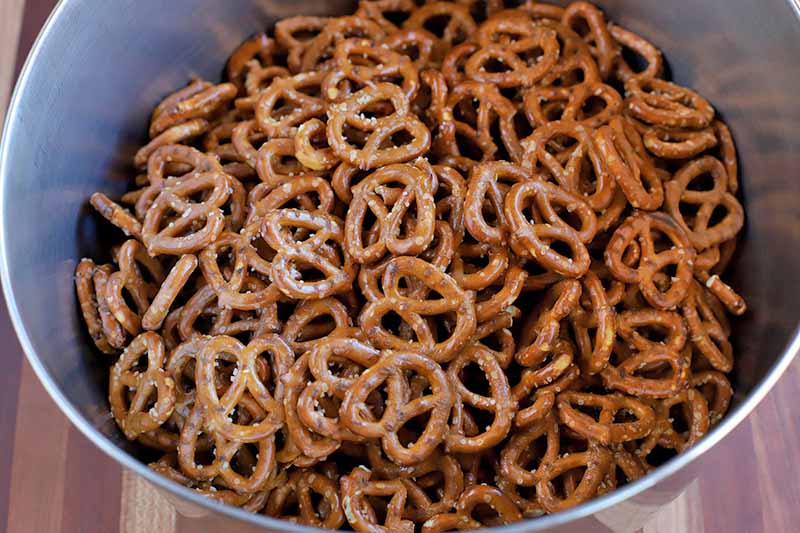 A large stainless steel bowl full of small, brown, store-bought salted pretzels, on a light brown wood surface.