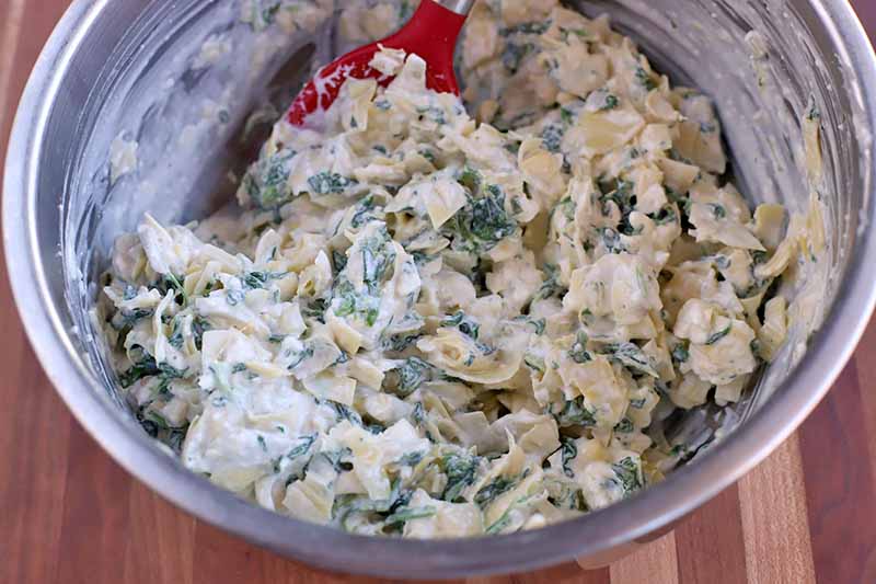 A red plastic utensil stirs a mixture of mayonnaise, artichoke hearts, and spinach, in a large stainless steel mixing bowl on a striped brown wood surface.