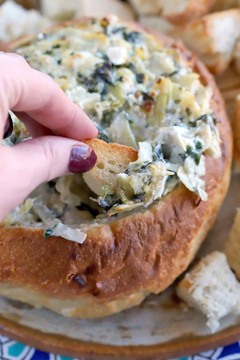 A manicured hand with burgundy nails dips a piece of bread into homemade spinach artichoke dip, in a hollowed out boule loaf, on a plate.