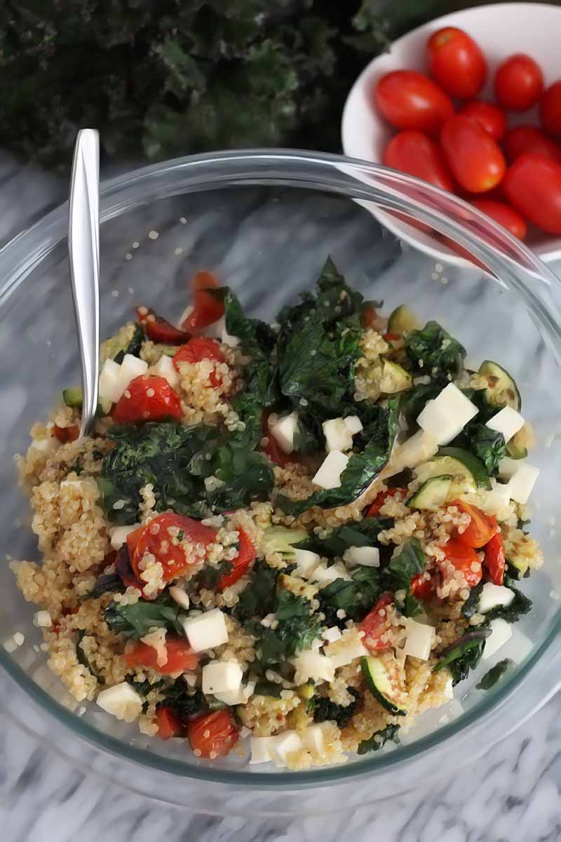 Overhead vertical shot of a glass mixing bowl of kale, quinoa, and vegetables with a spoon, on a gray marble surface with kale and a small white bowl of tomatoes in the background.