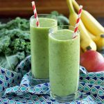 Two tall glasses filled with a green smoothie with red and white striped straws are at the center of the frame, surrounded by kale, bananas, and apples on a dark and light blue patterned cloth, on a dark brown surface with a lighter brown background.
