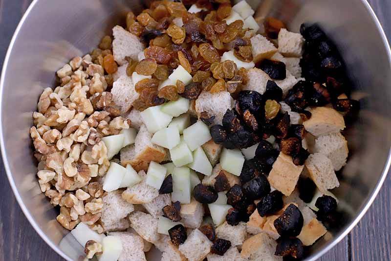 Overhead closely cropped shot of chopped apple, golden raisins, chopped dried figs, walnut pieces, and chunks of whole wheat bread in a stainless steel mixing bowl on a dark brown wood background.