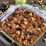 A square glass baking dish with handle cutouts on either side is filled with whole wheat bread pudding, on top of a white cloth kitchen towel with scattered walnut pieces, raisins, dried figs, and two green apples, on a dark brown wood surface.