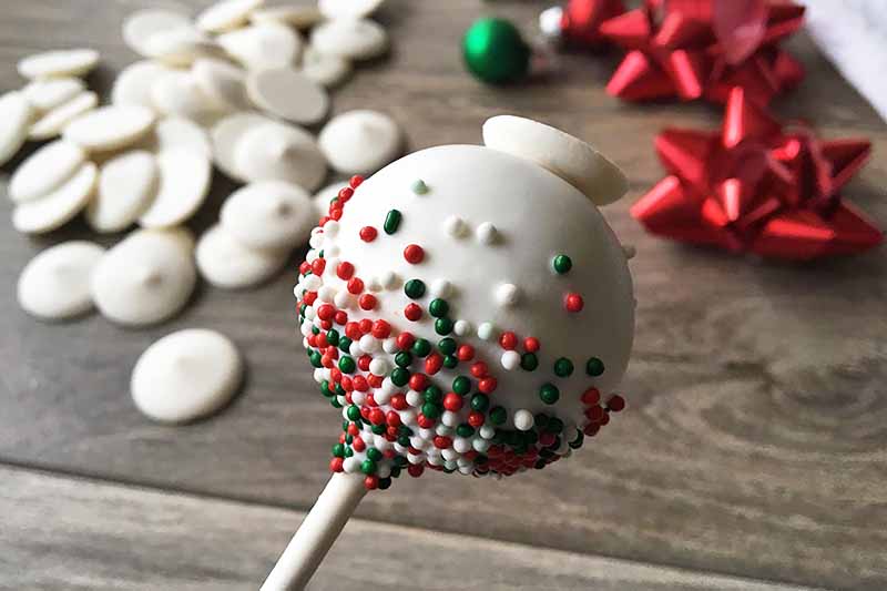 Horizontal image of a decorated white cake pop with sprinkles and white chocolate wafers and decorations in the background.