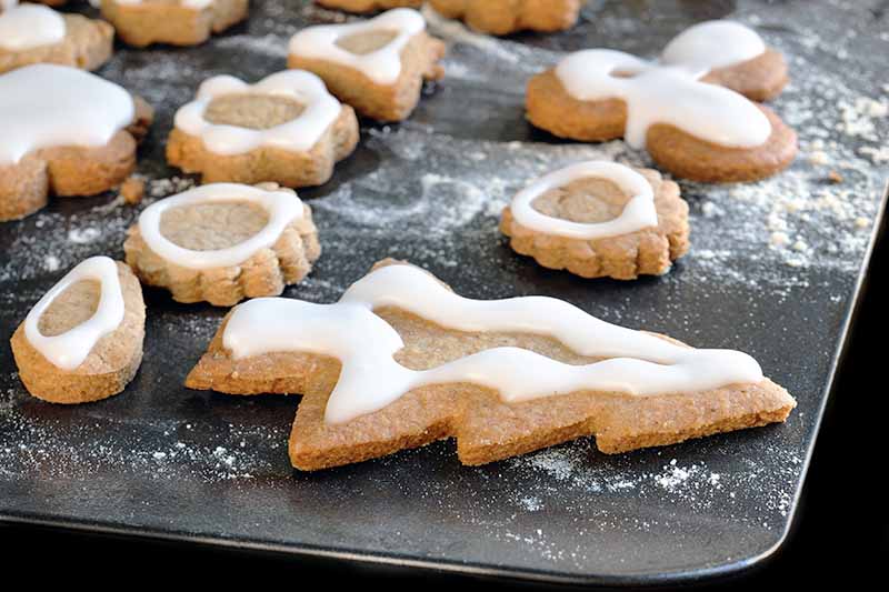 Closeup of cutout Christmas cookies decorated with white sugar icing on a rimless metal baking sheet, on a black background.
