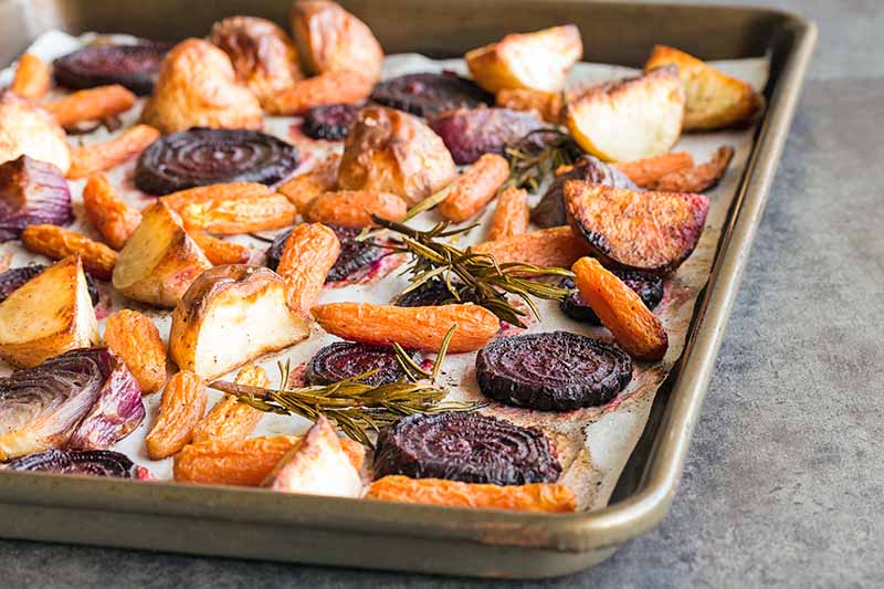 Closeup closely cropped horizontal image of a rimmed metal sheet pan lined with parchment paper and filled with slice droasted carrots, beets, and other vegetables with sprigs of rosemary, on a gray speckled countertop.