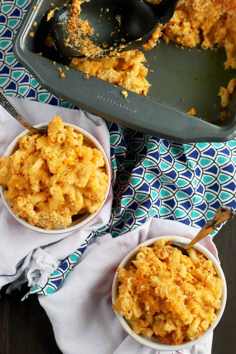 Overhead vertical shot of two dishes of mac and cheese with forks stuck into them, with a metal baking pan of more of the casserole with a black plastic serving spoon, on top of two folded white cloths and a wrinkled dark and light blue patterned cloth, on a dark brown wood surface.