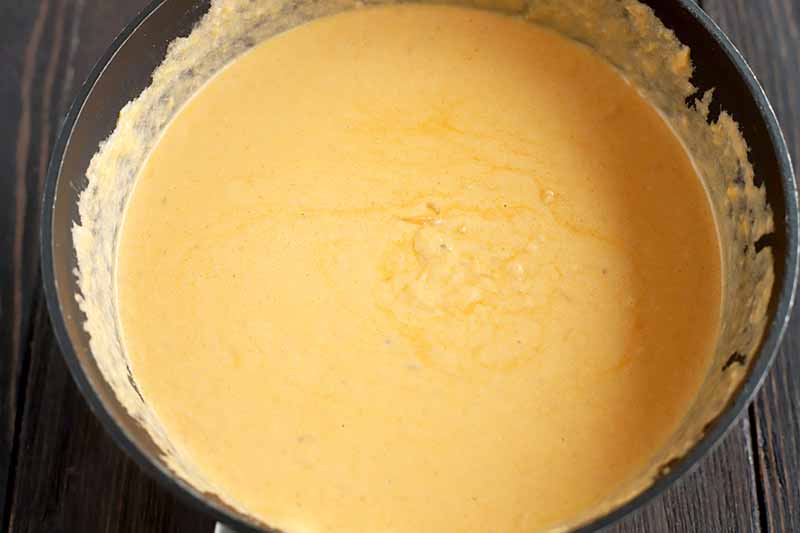 Overhead closely cropped shot of a homemade cheese sauce in a nonstick saucepan, on a dark brown wood table.