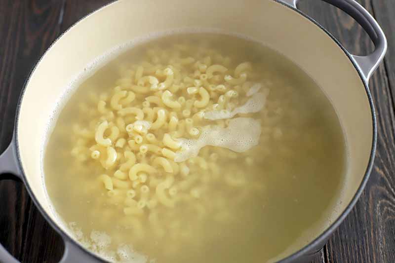 Cooked elbow macaroni in a large enameled pot filled with water, on a dark brown wood surface.