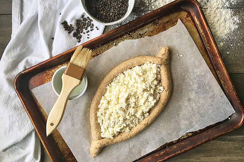 Horizontal image of unbaked shaped dough filled with white ingredients on a baking pan next to a pastry bush and bowl of peppercorns.