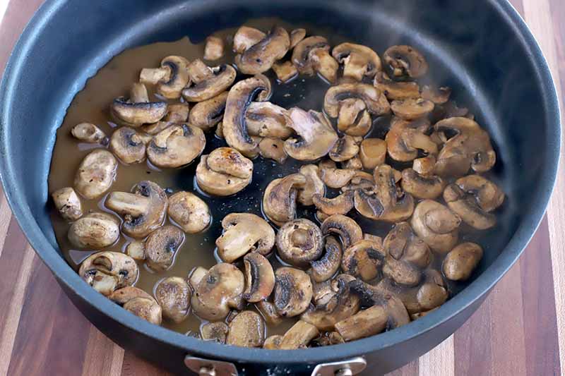 Sauteed sliced mushroom in a large nonstick frying pan with chicken broth and marsala wine, on a wood surface.