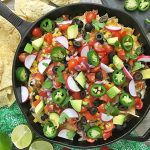 Horizontal image of a cast iron skillet with various nacho toppings next to chips on a green napkin