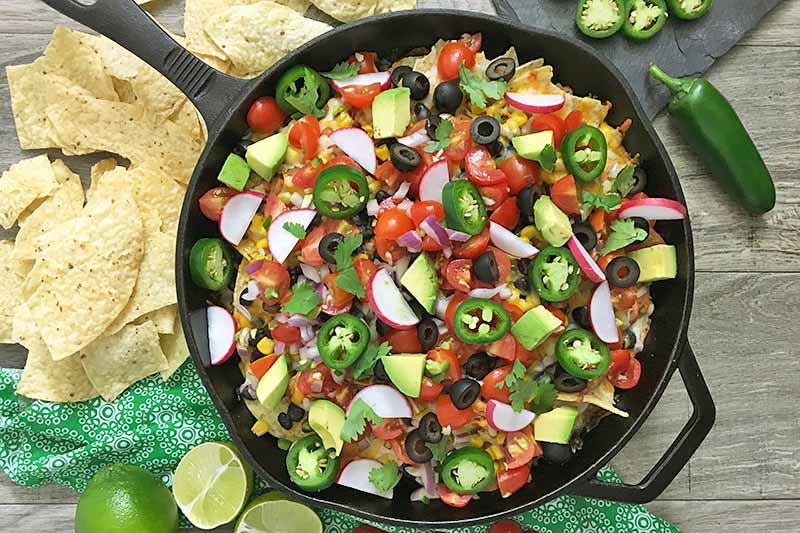 Horizontal image of a cast iron skillet with various nacho toppings next to chips on a green napkin