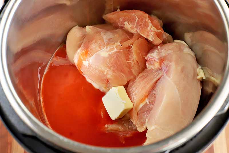 Raw chicken breast, a pat of butter, and hot sauce in a metal and black plastic slow cooker, on a wood surface.